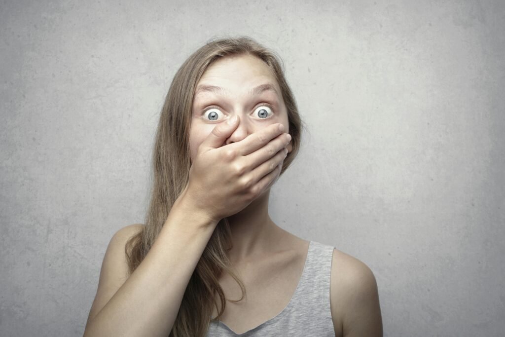 A startled woman covers her mouth expressing surprise against a neutral background.