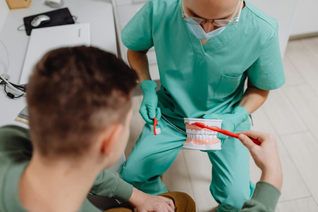 A dentist in green scrubs shows a patient how to brush teeth using a dental model.