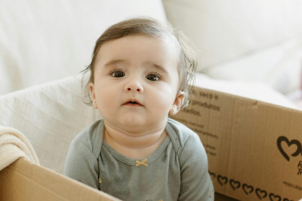 pexels-photo-26509769-26509769 Charming portrait of a baby sitting in a cardboard box indoors, captured with soft lighting.